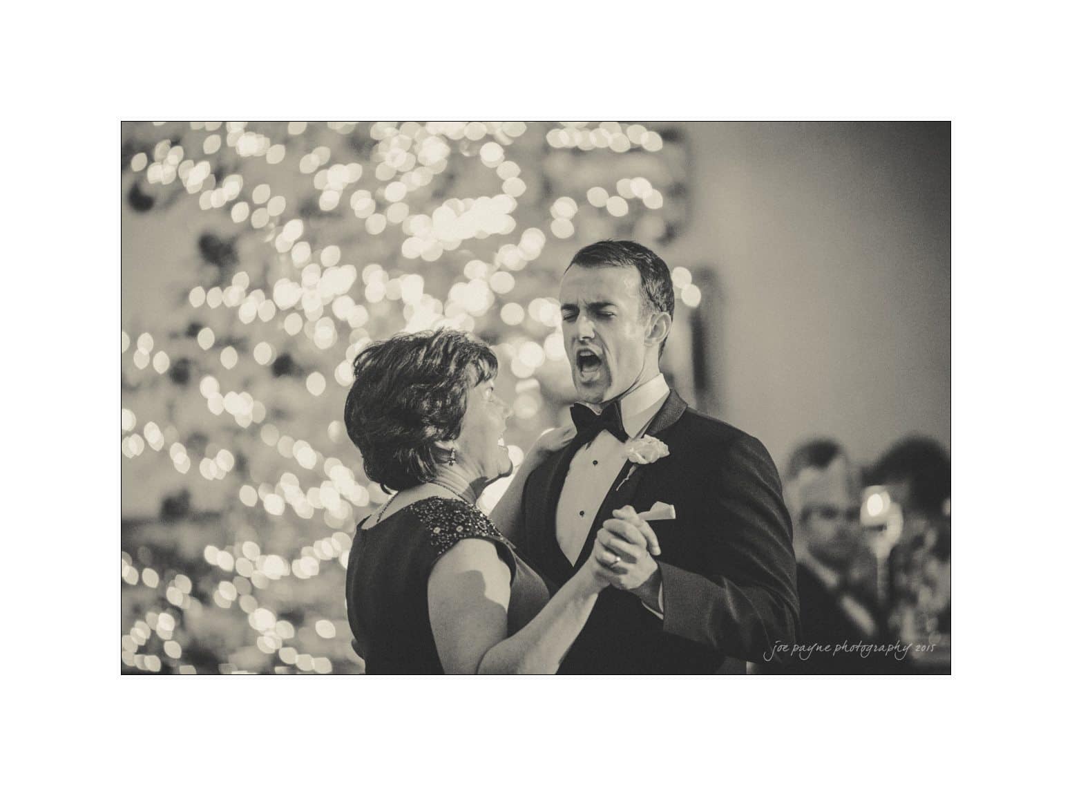 A man and a woman, dressed formally, dance together. The man appears to be singing or speaking expressively. In the background, soft lights create a bokeh effect. The scene is set in a warm, intimate atmosphere.