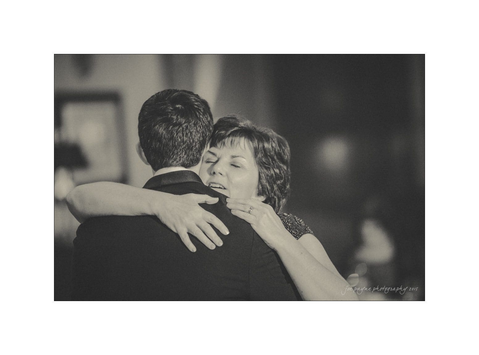 A monochrome photo of two people embracing closely. One person is facing the camera with a content expression, while the other has their back to the viewer. The background is softly blurred, creating a warm and intimate atmosphere.