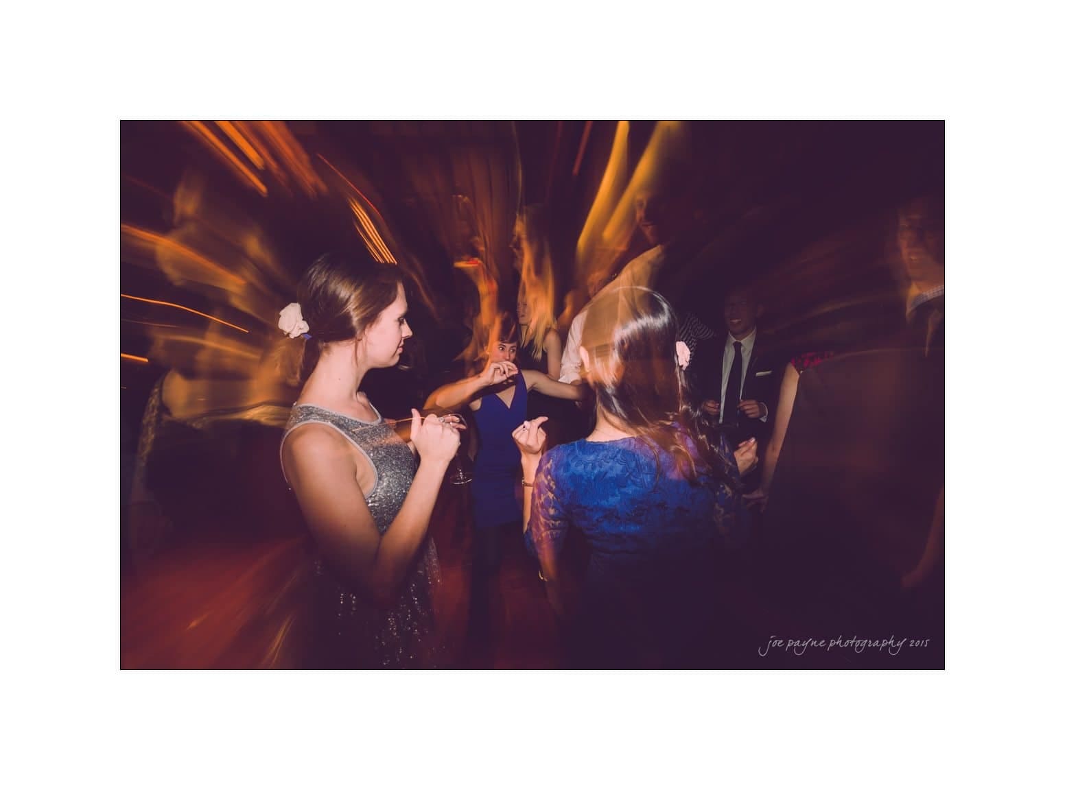 A group of people dancing in a dimly lit room with blurred lights creating a dynamic effect. The scene captures motion and energy, with individuals in formal attire appearing joyful and engaged in the moment.