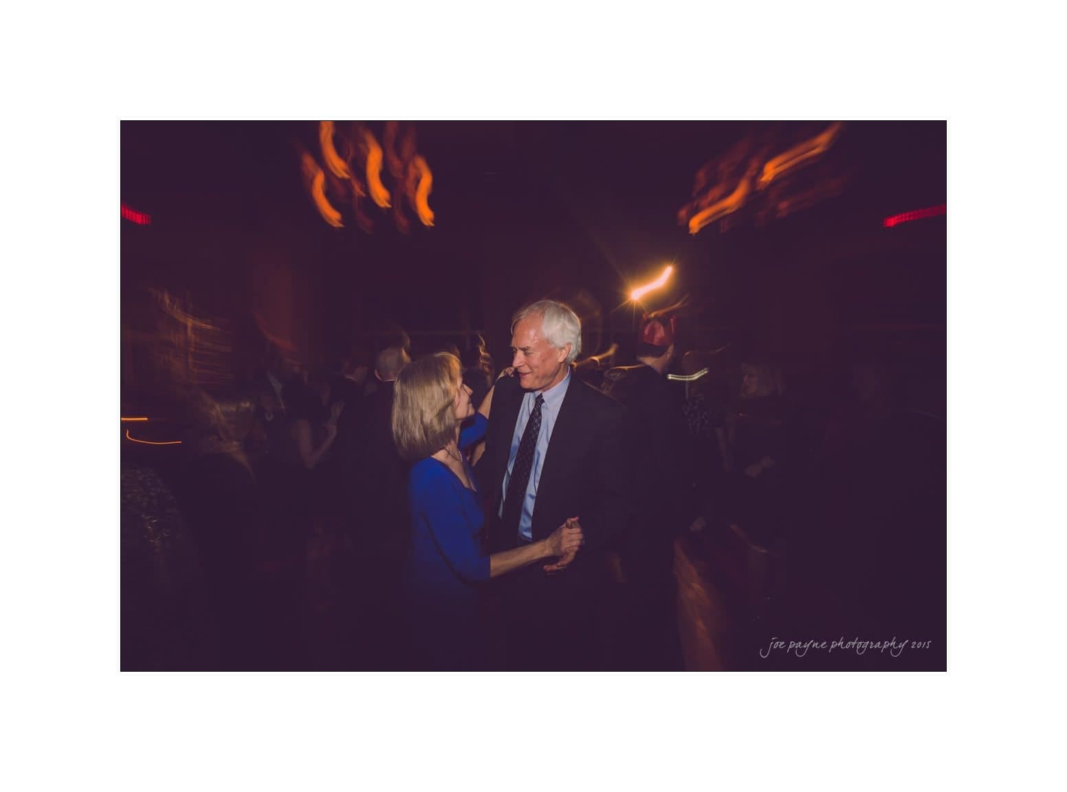 An older couple dances closely on a dimly lit dance floor, surrounded by blurred figures and soft, colorful lights in the background. The ambiance is warm and intimate.