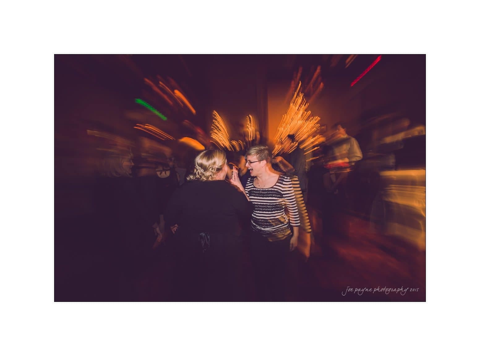 A candid photo of two women talking and laughing at a party. The background is blurred with warm lights and other indistinct people, creating a dynamic and lively atmosphere.