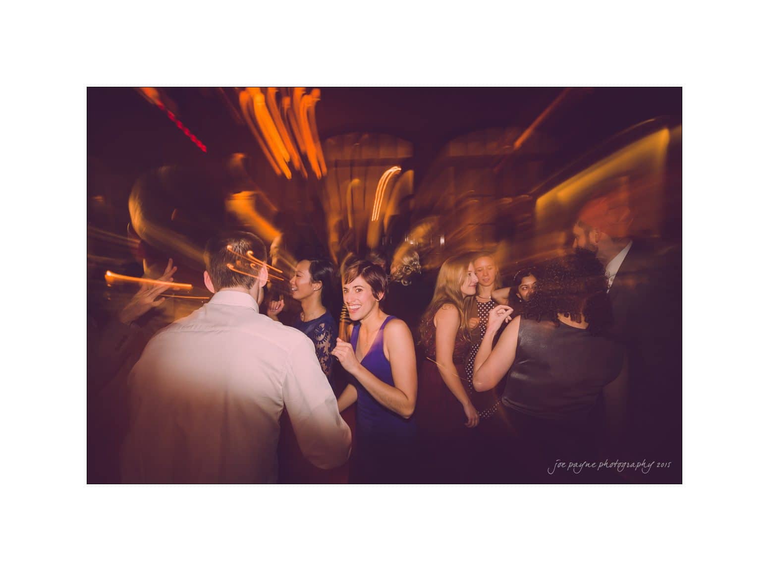 A group of people dancing energetically in a dimly lit room with a blurred effect. The scene captures motion and joy, with warm lighting casting a festive atmosphere.