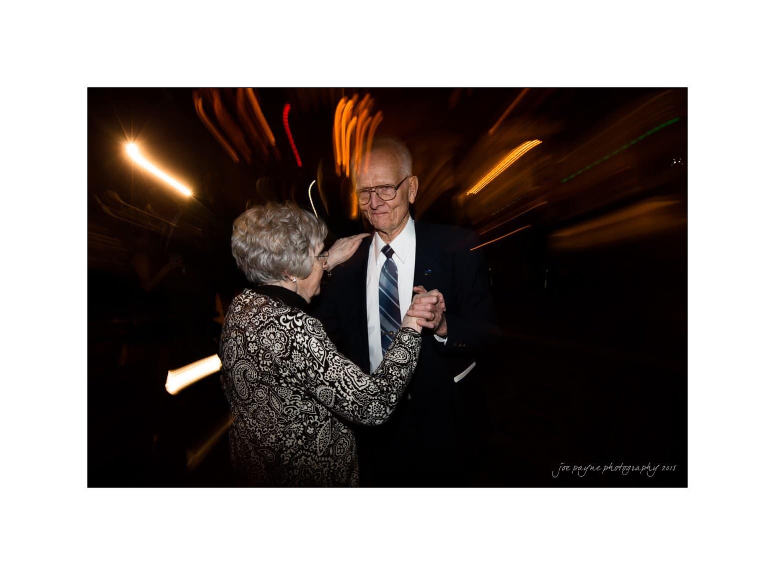 An elderly couple dances in low light, surrounded by blurred streaks of light creating a dynamic background. The man wears a suit and glasses, while the woman wears a patterned outfit. They smile at each other, captured in a moment of joyful connection.