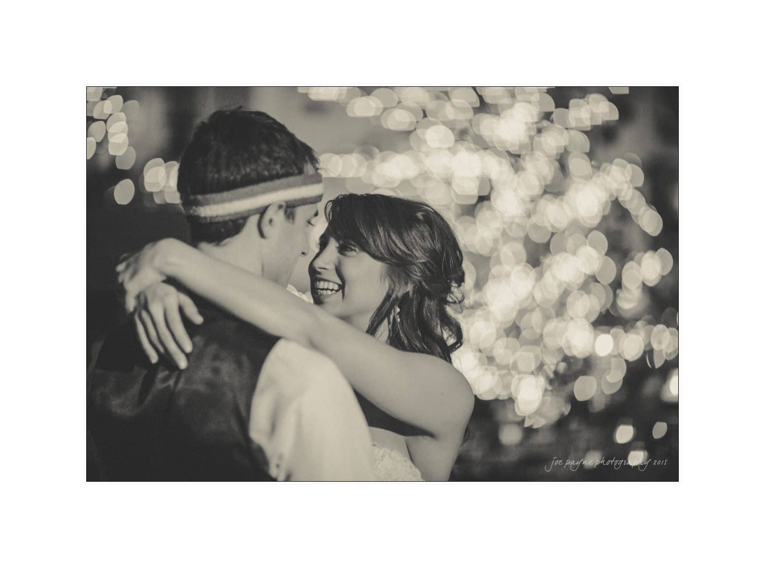 A black and white photo of a couple embracing and smiling at each other, surrounded by softly lit bokeh lights. The woman is wearing a strapless dress, and the man has a headband and vest.