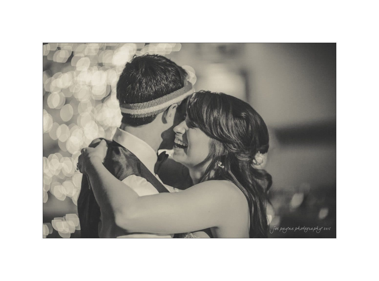 A black and white photo shows a couple embracing and dancing closely. The woman, smiling, rests her head near the mans shoulder. The background is softly blurred, creating a romantic, intimate atmosphere.