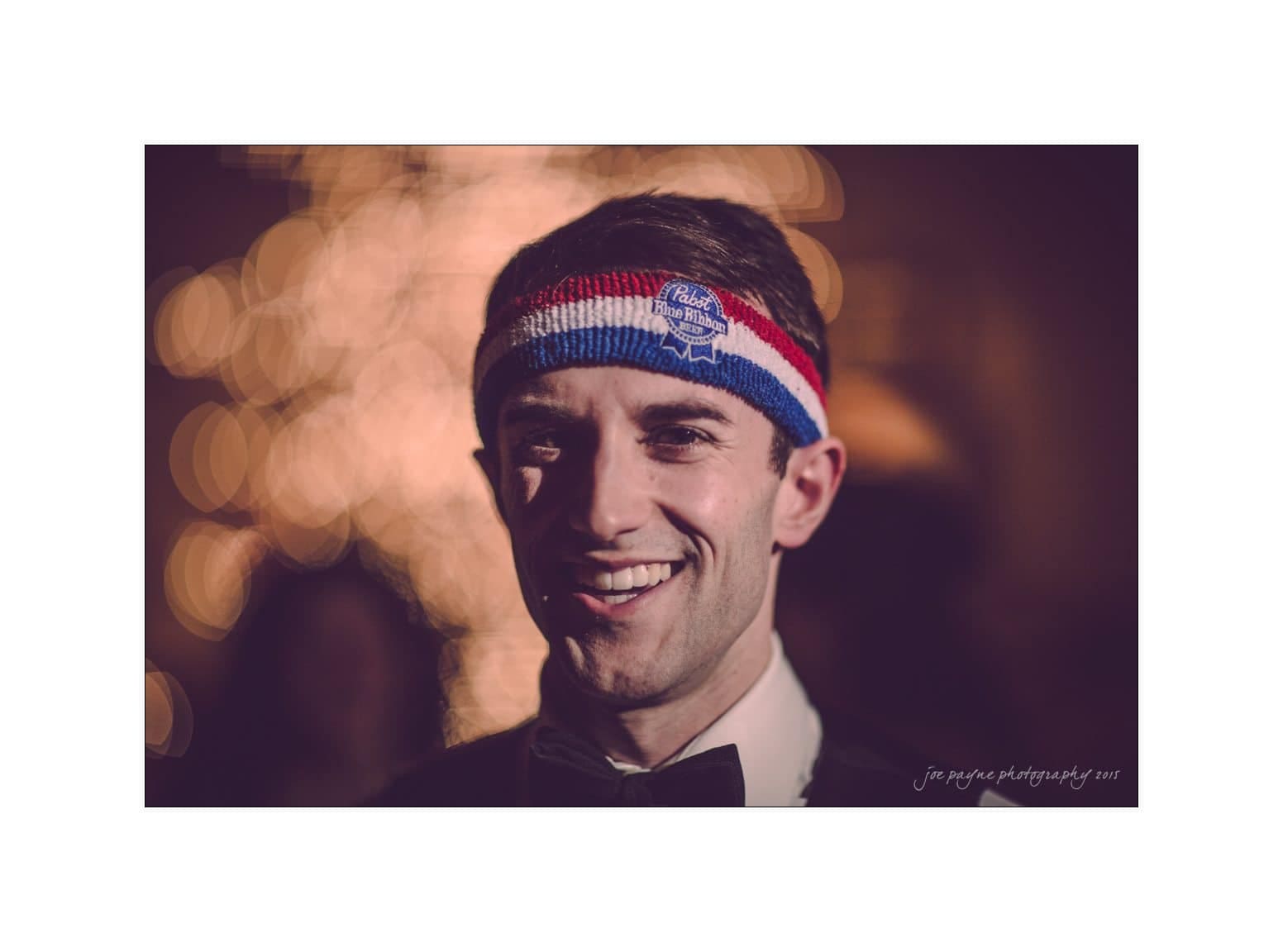A man smiling at the camera, wearing a dark suit, bow tie, and a colorful headband. The background is softly blurred with bright lights.