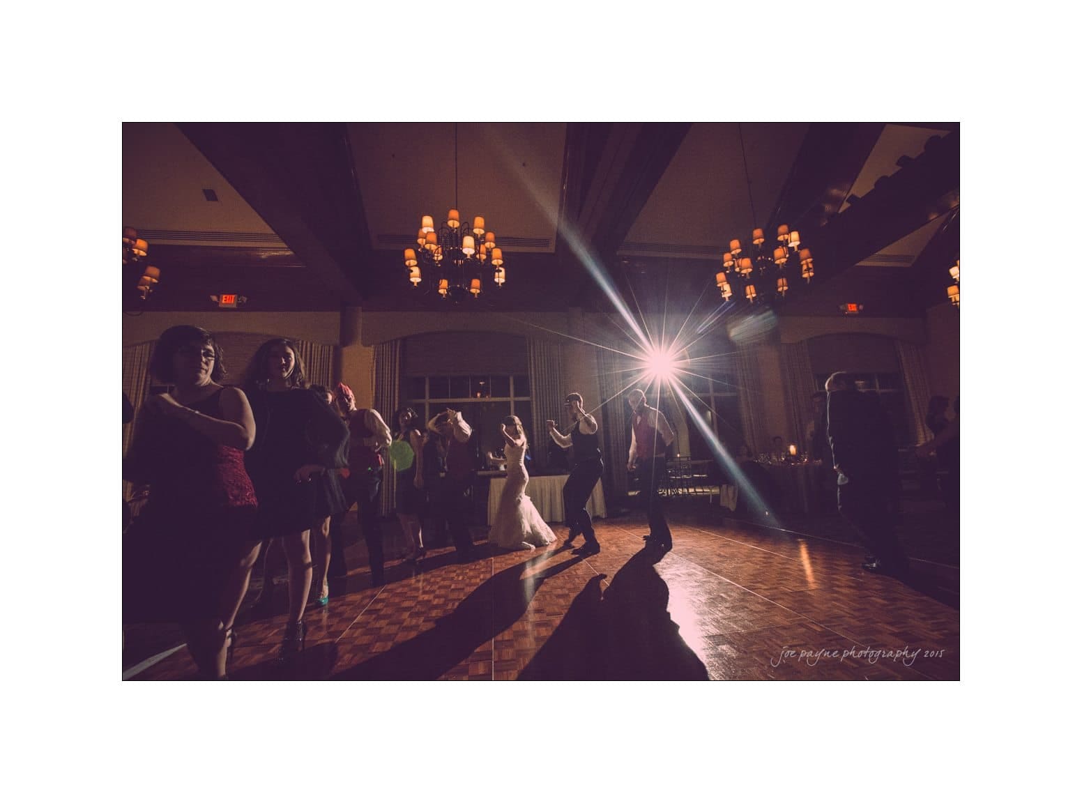 People are dancing in a warmly lit hall with a polished wooden floor. The scene features soft, ambient lighting from chandeliers above, with a bright spotlight illuminating the dancers from behind, casting long shadows.