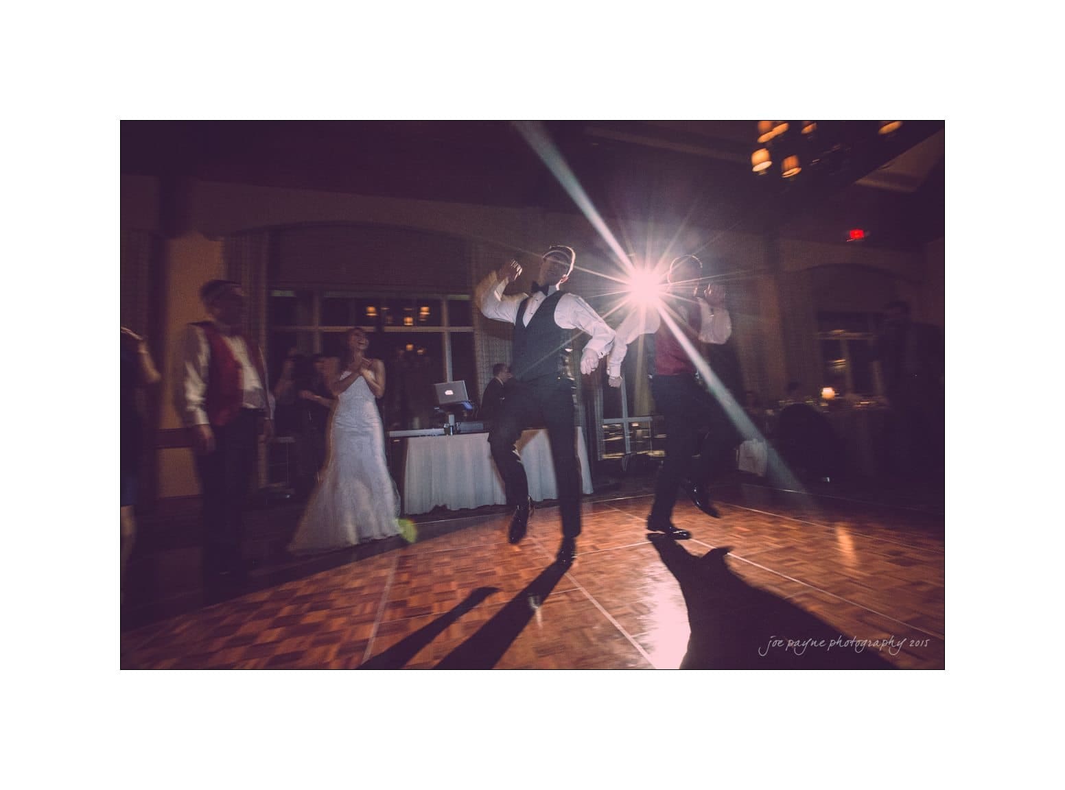 A bride and groom dance energetically on a wooden floor at a wedding reception. The groom is captured mid-jump, illuminated by a bright spotlight in the background. Guests in formal attire are visible in the dimly lit room.