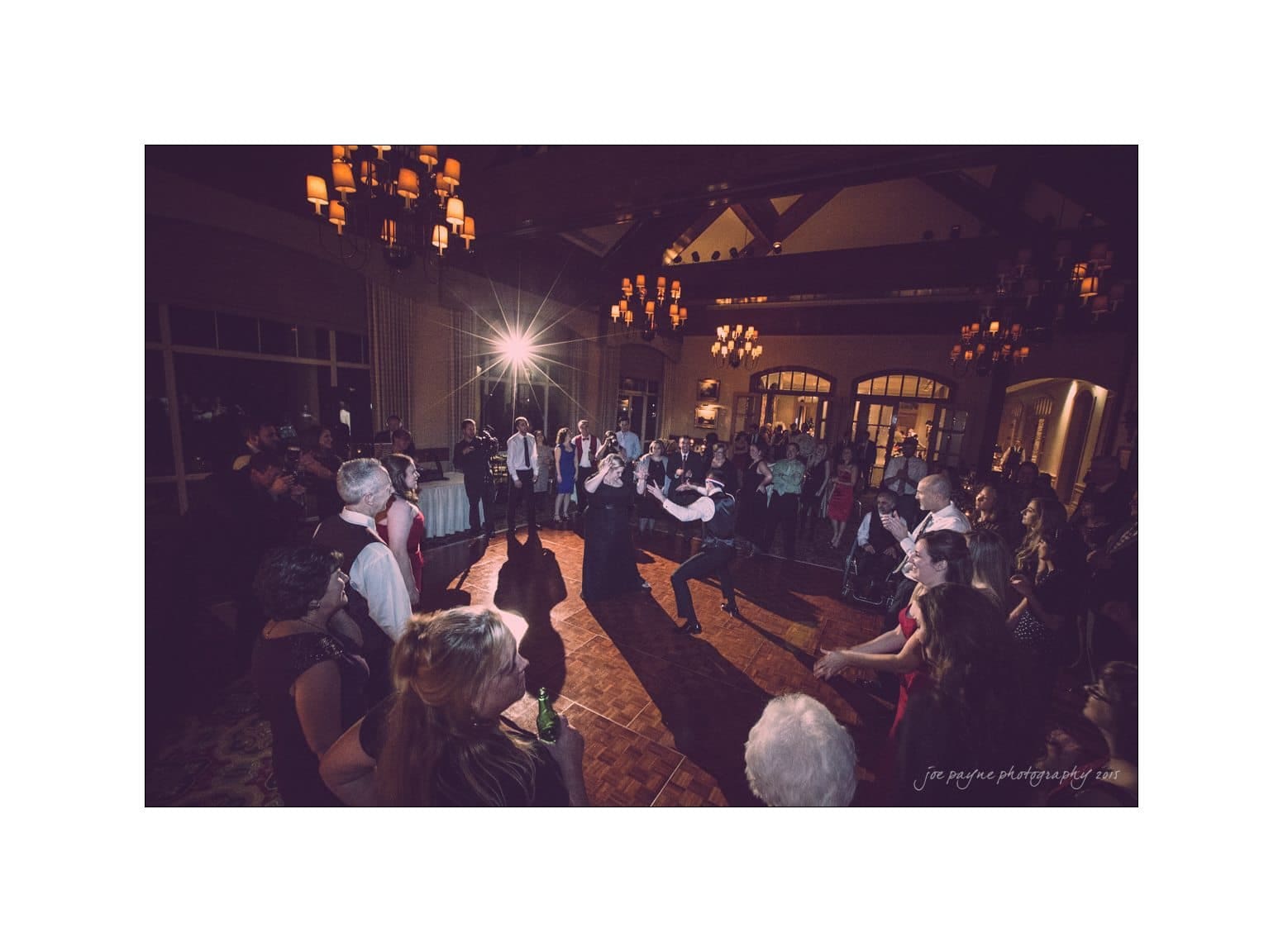 A lively dance floor at a wedding reception with a couple twirling in the center. Guests form a circle around them, some clapping and watching with joy. The venue is warmly lit with chandeliers, creating a festive and elegant atmosphere.