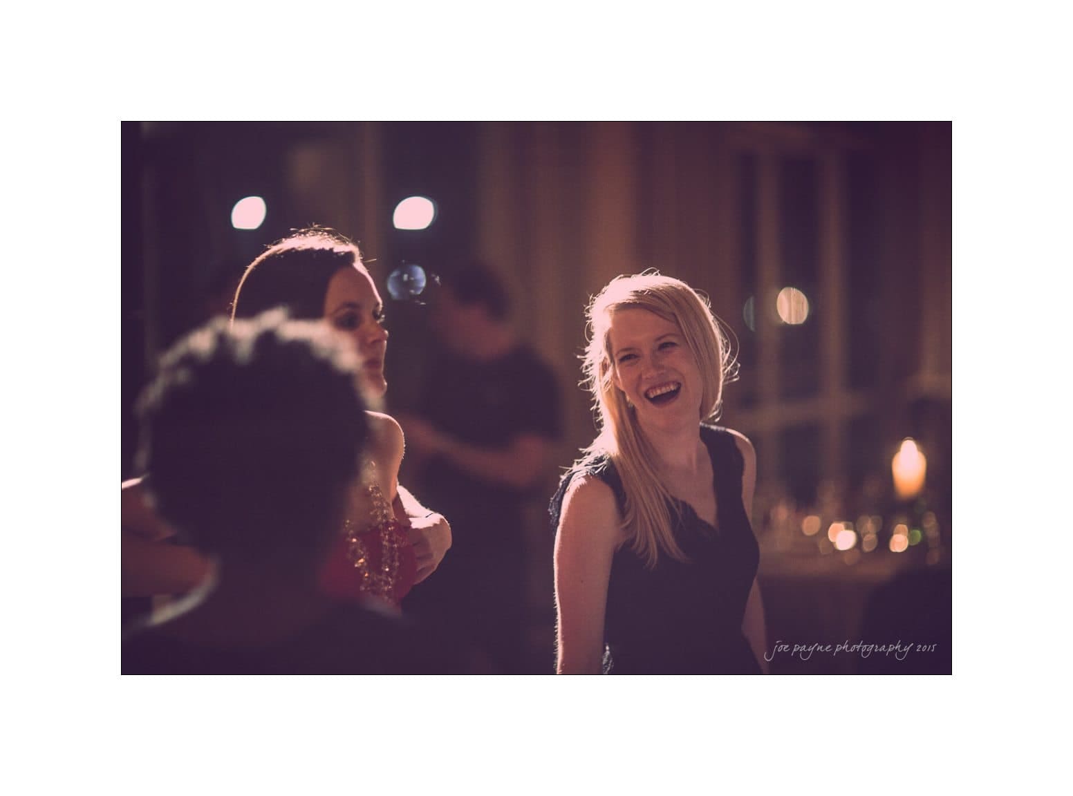 A group of people in formal attire enjoy a lively atmosphere at an indoor event. The focus is on a smiling woman with long hair, wearing a dark dress. Blurred lights create a warm, festive ambiance in the background.