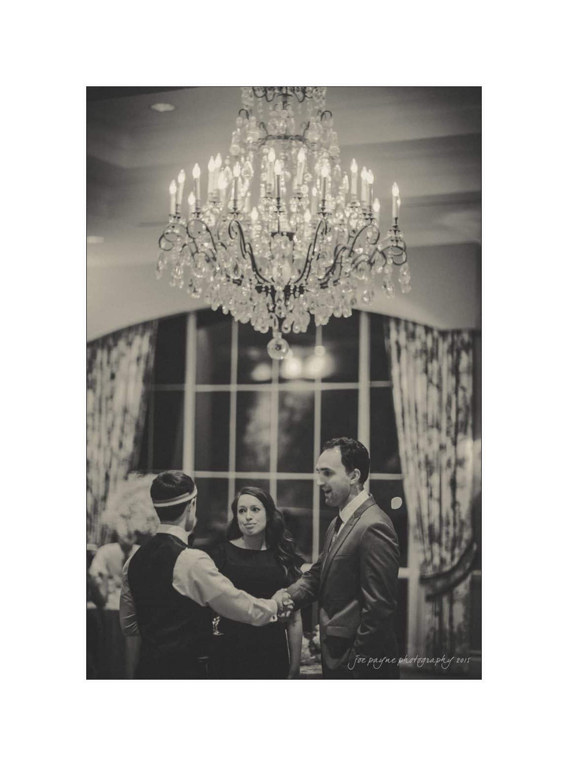 A black-and-white image of two men shaking hands under a large chandelier. A woman stands between them, looking on. The backdrop includes a window with draped curtains.