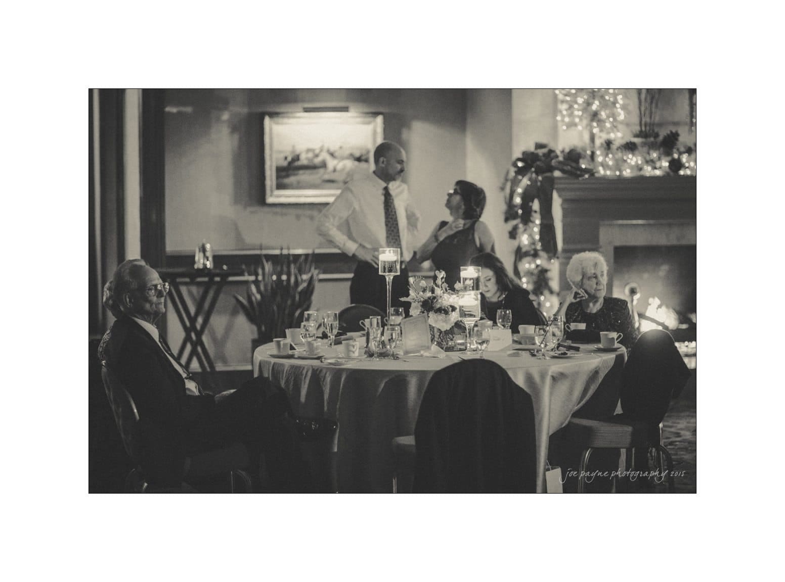 A black and white photo of a dinner party. Four people sit around a table with empty glasses and plates, engaged in conversation. A man and woman stand near a fireplace adorned with garlands and lights. The atmosphere is warm and intimate.