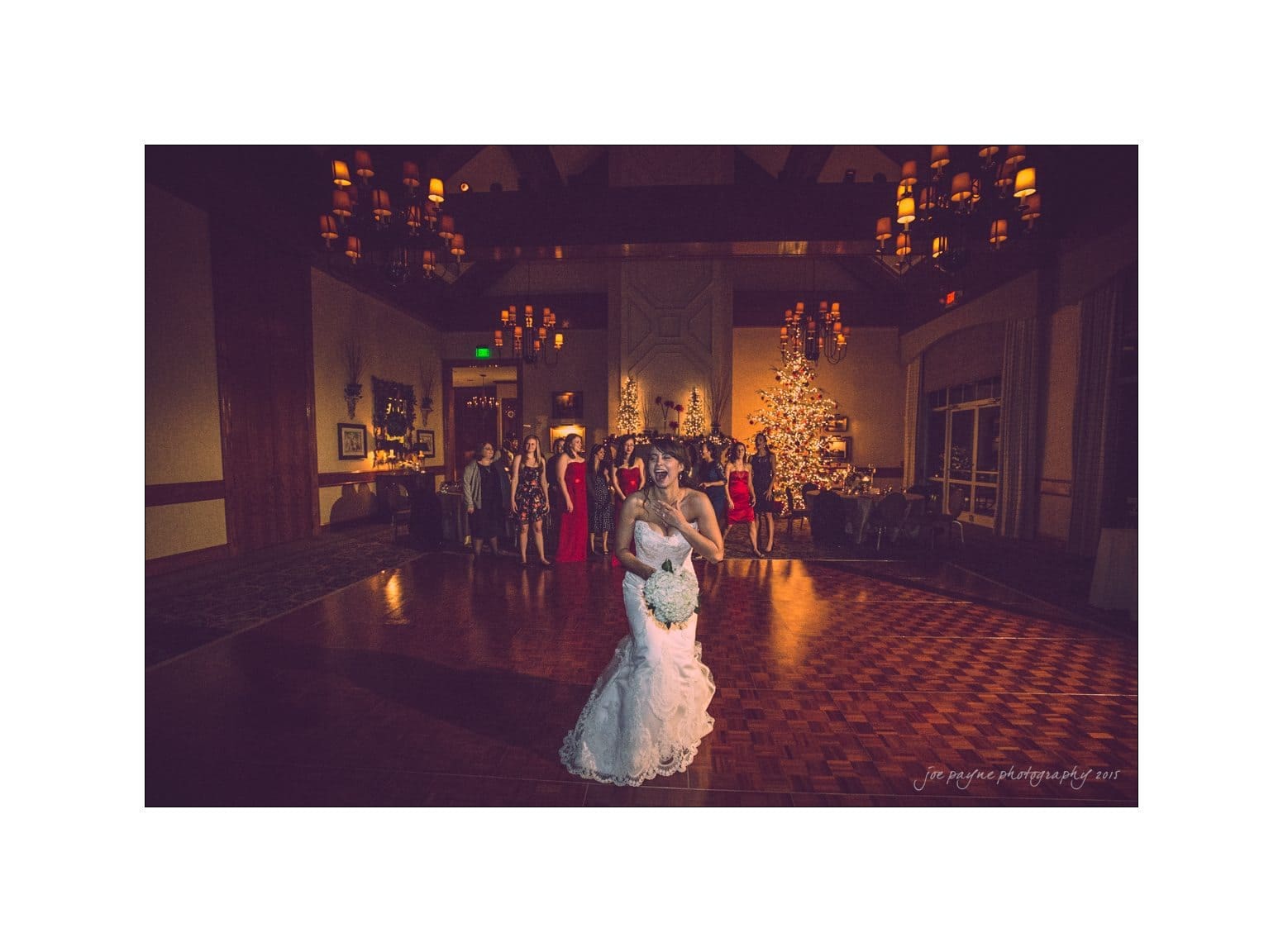 A bride stands on a dimly lit dance floor holding a bouquet. In the background, a group of women anticipate the bouquet toss. The room is warmly lit with chandeliers and a decorated Christmas tree.