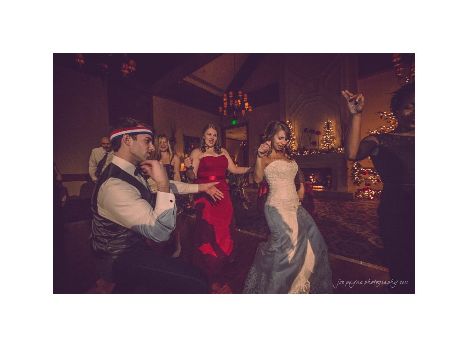A group of people in formal attire dance energetically at a festive, warmly lit event. A man in a vest and headband gestures playfully, while a woman in a red dress and another in a white gown join in, with a decorated Christmas tree in the background.