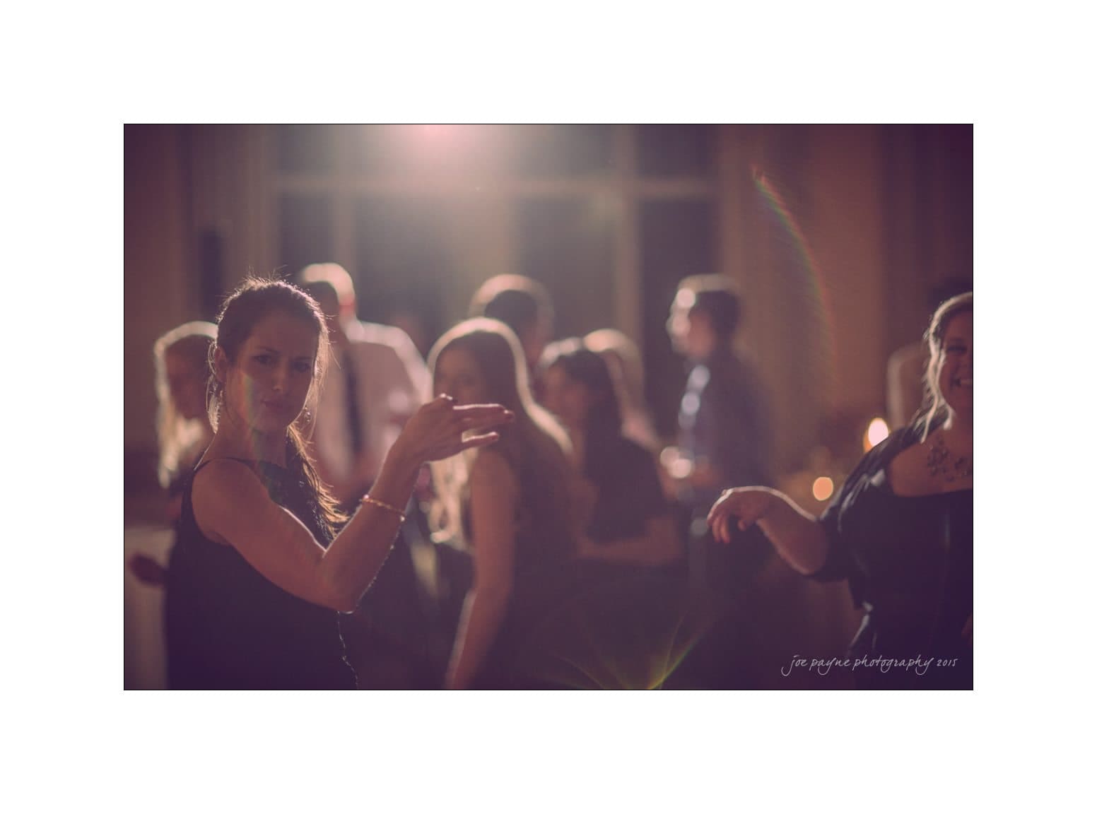 A group of people are gathered at a dimly lit indoor event. The focus is on a woman gesturing playfully towards the camera. The background shows others mingling and softly illuminated by ambient lighting.