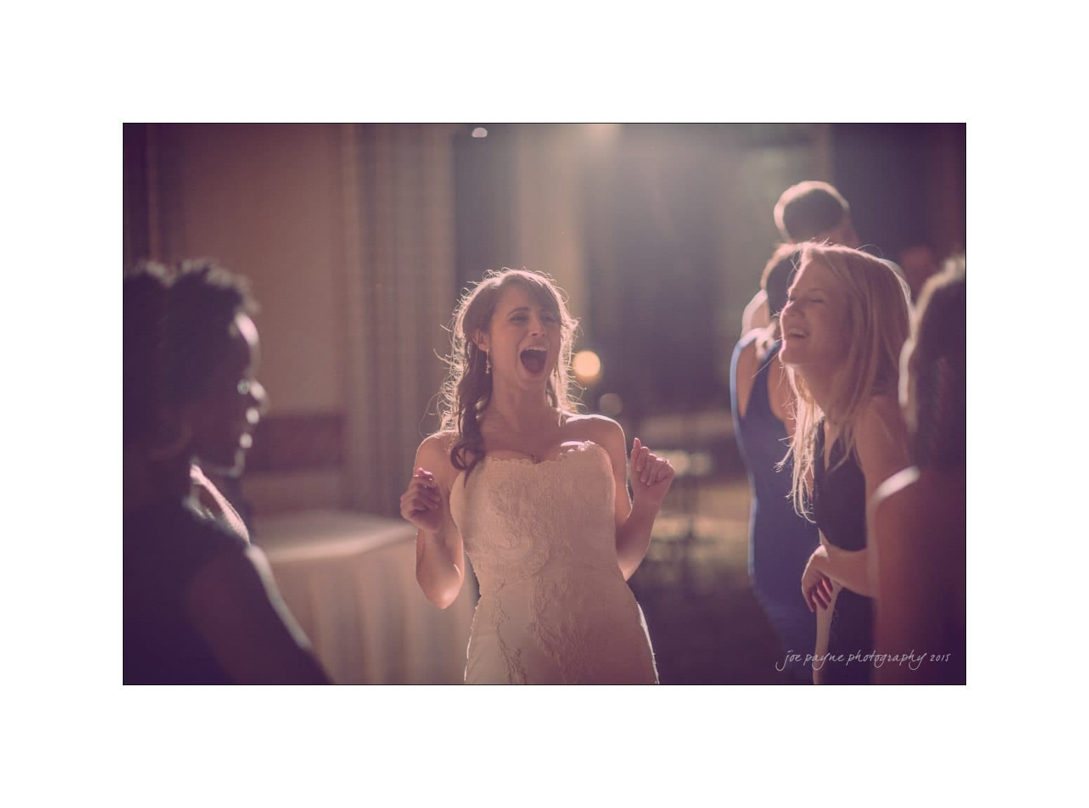 A bride in a white dress joyfully dances and laughs with friends at a wedding reception, set in a softly lit room. The atmosphere is festive and filled with smiles and movement.