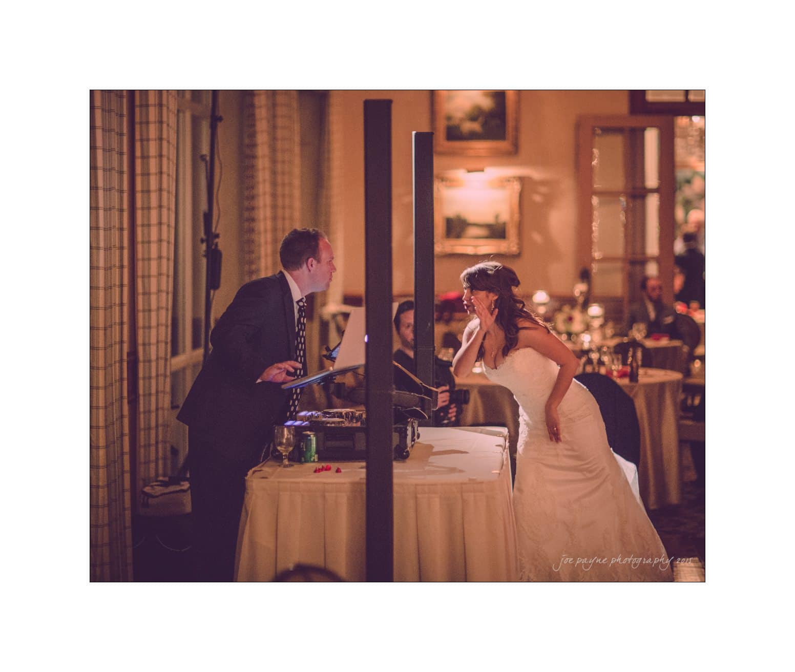 A bride in a wedding dress playfully interacts with a DJ at a reception. The setting is warmly lit, with an elegant backdrop featuring framed pictures and dining tables. The mood is joyful and celebratory.