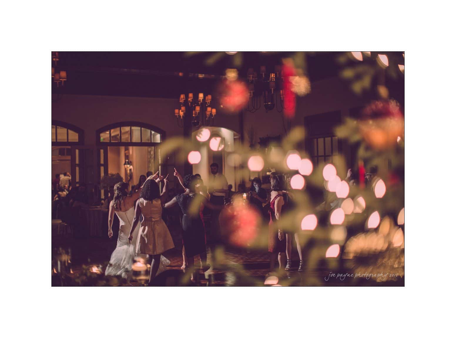 A group of people are dancing in a warmly lit room, with soft, blurred lights and festive decorations in the foreground creating a cozy, celebratory atmosphere.