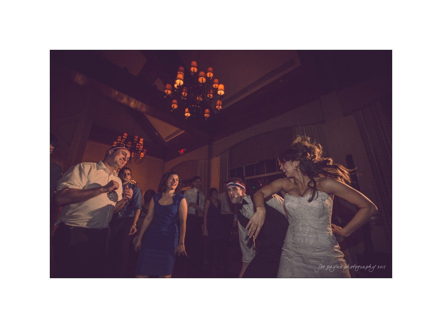 A group of people dance at an indoor event under a chandelier. A woman in a light-colored dress leads the dance, with others joyfully following her movements. The dim lighting adds a warm, festive atmosphere to the scene.
