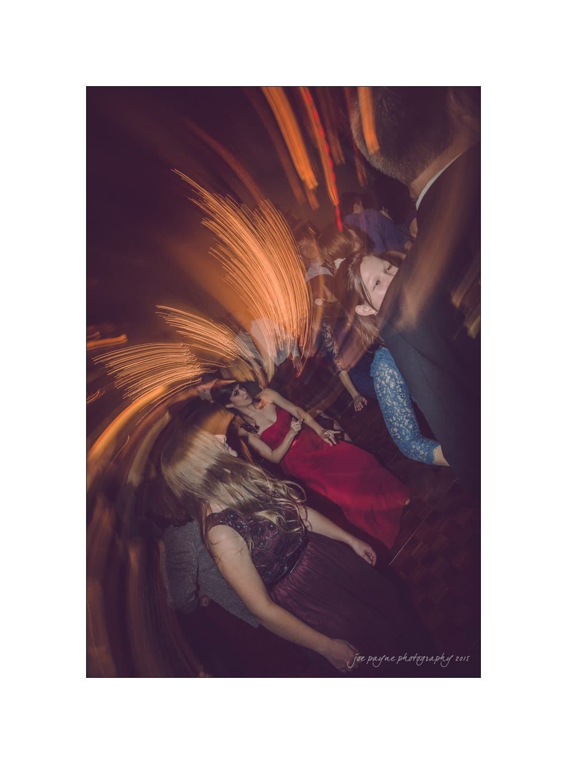 A group of people dancing at an event with motion blur effects. One person in a red dress is prominent in the background. Warm lighting creates streaks, adding a dynamic and energetic atmosphere to the scene.
