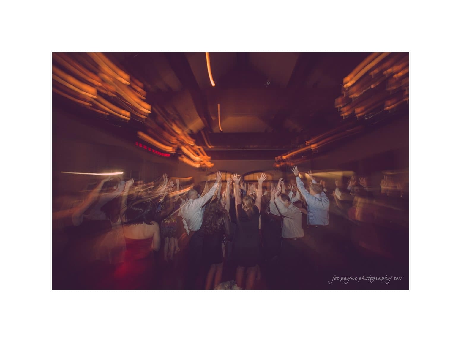 A crowd of people dancing energetically in a dimly lit room with blurred motion, capturing the lively atmosphere. The ceiling lights create streaks of light, enhancing the dynamic feeling of the scene.