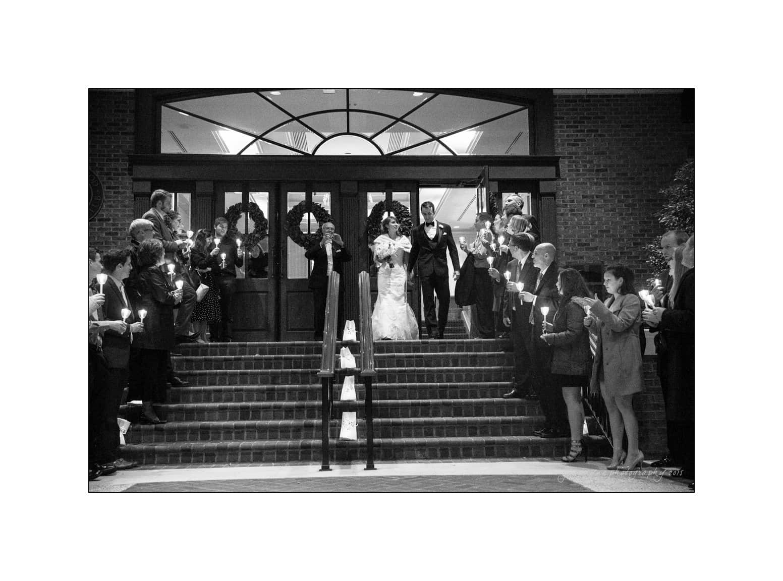 A bride and groom exit a building, walking down steps lined with lit candles. They are surrounded by guests holding candles, creating a warm and celebratory atmosphere. The setting appears to be at night, adding to the intimate ambiance.