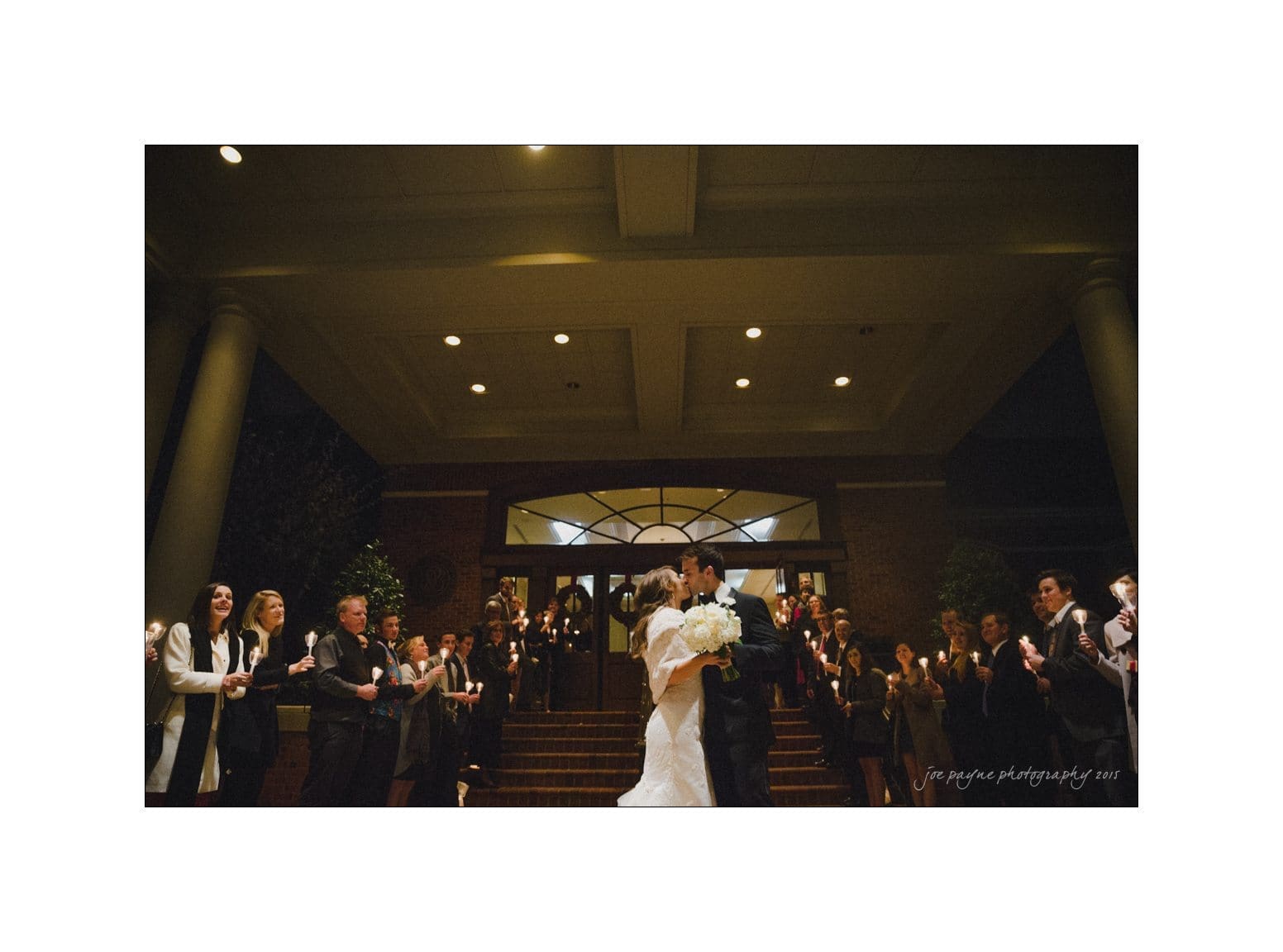 A bride and groom share a kiss at the top of a staircase, surrounded by cheering friends and family at night. The guests hold sparklers as they celebrate under a well-lit building entrance.