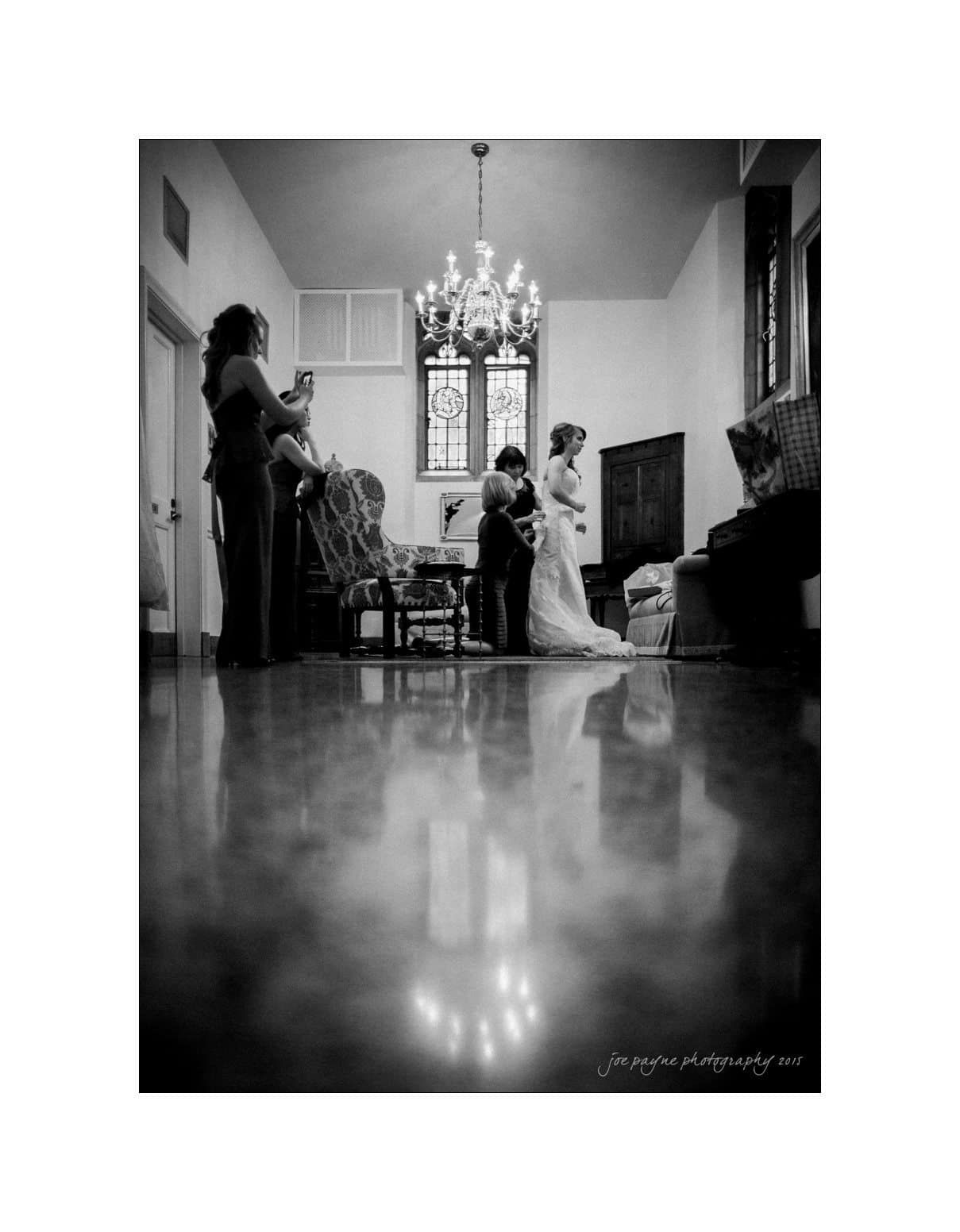 A black and white photo of a bride having her dress adjusted by a woman in a room with a large chandelier. Another woman takes a picture with her phone. The room has a reflective floor and a large, decorative window.