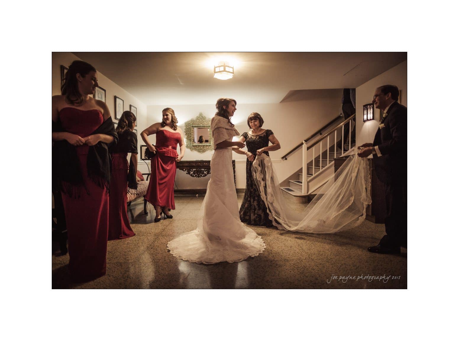 A bride in a white gown stands with four bridesmaids and an older man in a dimly lit room. The bridesmaids wear red dresses, and the man holds the brides veil. The scene seems calm and elegant, with soft lighting above.