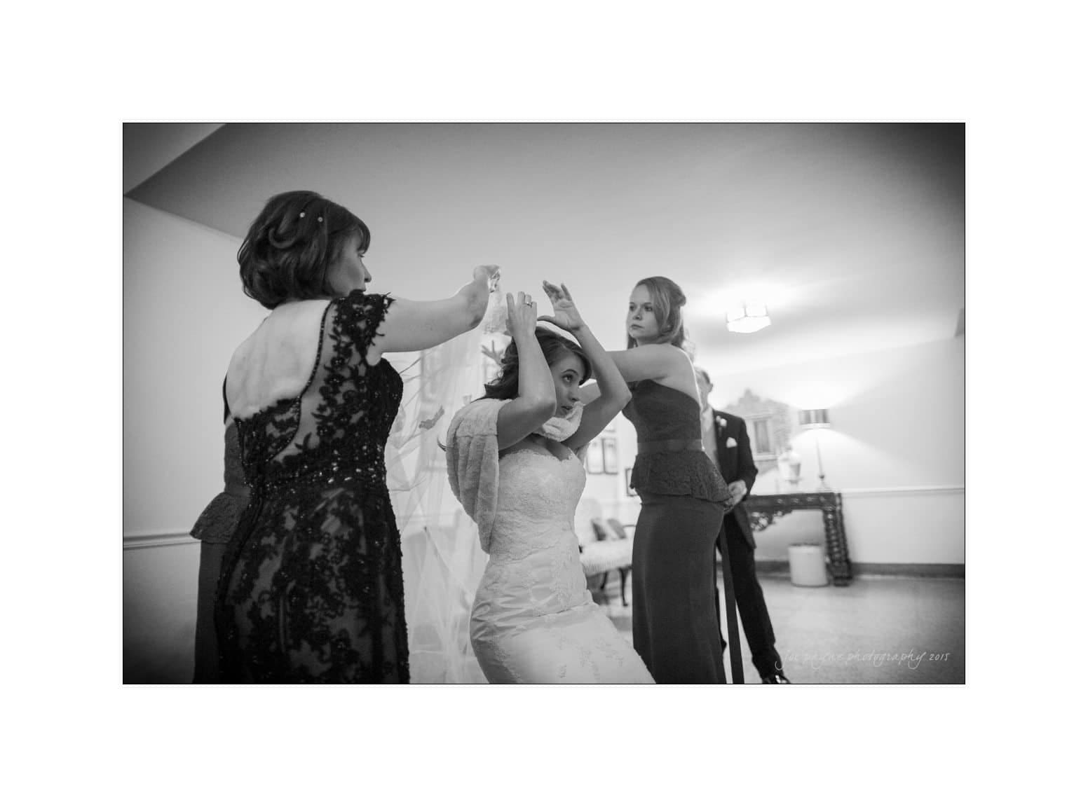 A bride in a lace gown adjusts her veil with help from two women, one wearing a dark lace dress. Another woman in the background looks on. The scene is set indoors with soft lighting, capturing a candid moment before a wedding.