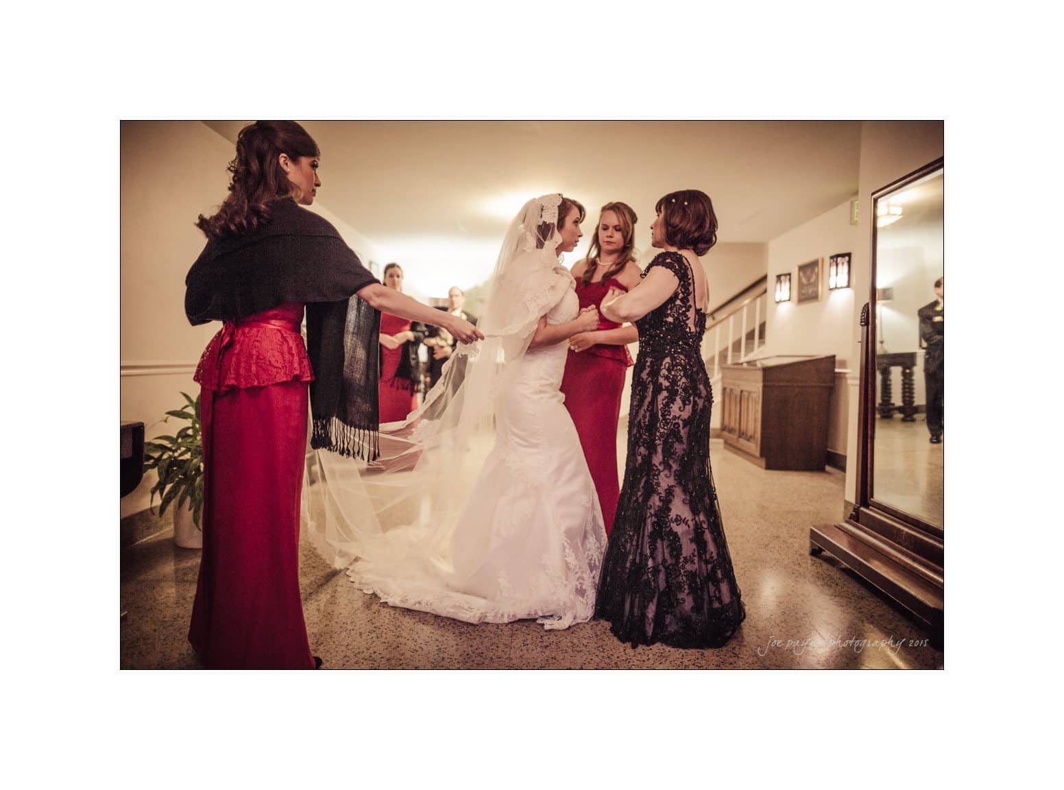 A bride in a white gown and veil is being helped by three women in elegant dresses. They are adjusting her train in a warmly lit room with a mirror and wooden flooring.