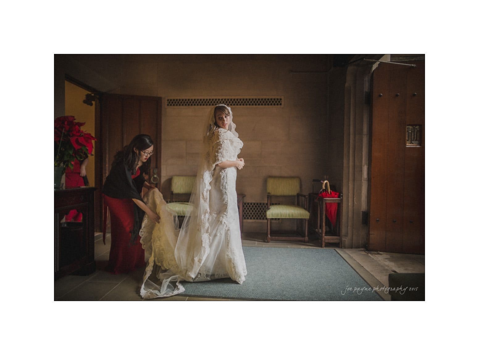 A bride in a wedding dress stands in a dimly lit room, adjusting her veil. Another woman in a red dress crouches behind her, arranging the train of the gown. Two chairs and a red umbrella are in the background.