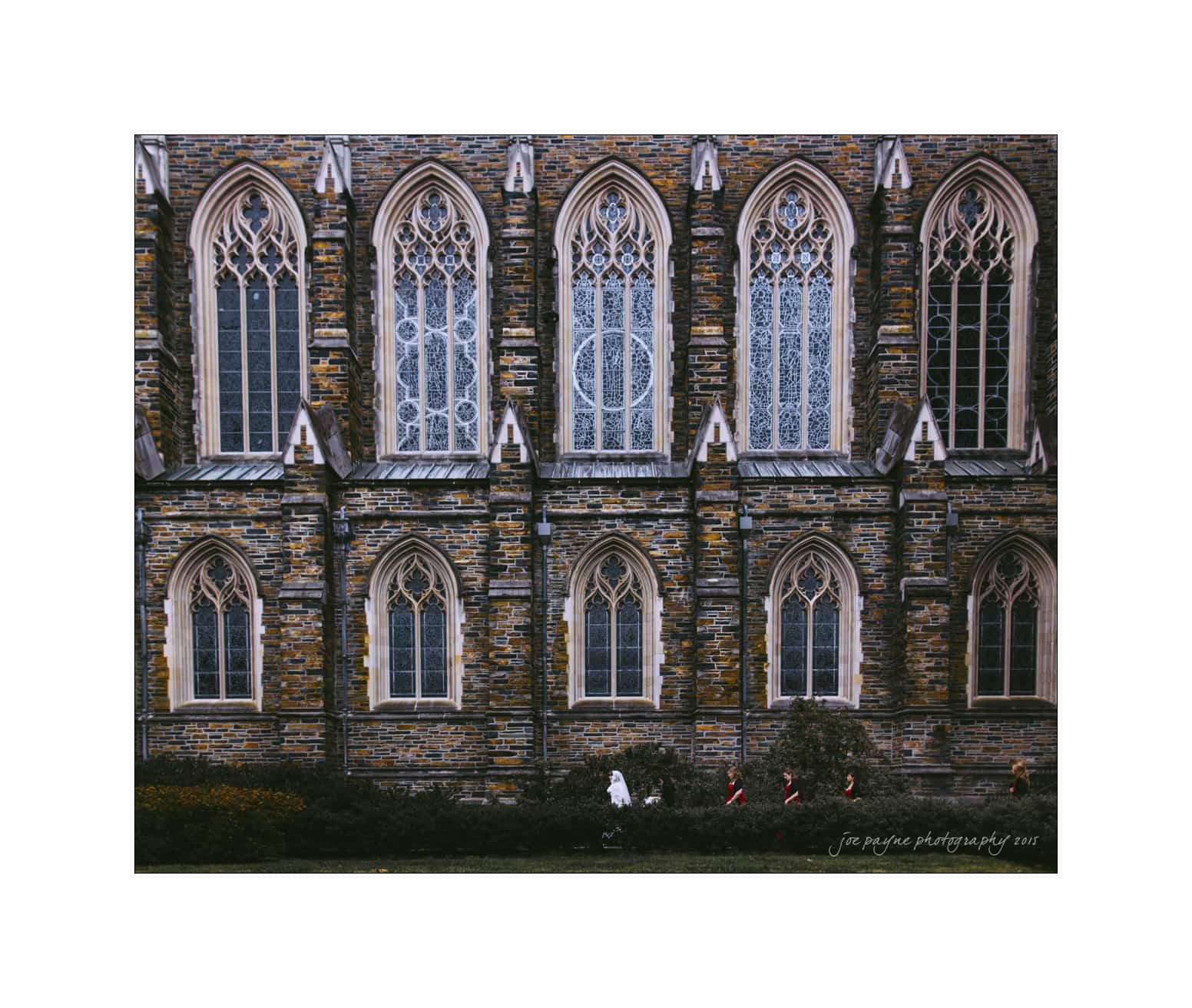 A stone building with six tall, narrow, arched windows lined with intricate designs. Three people walk along a path at the base of the building, surrounded by greenery. The photo has a signature in the bottom right corner.
