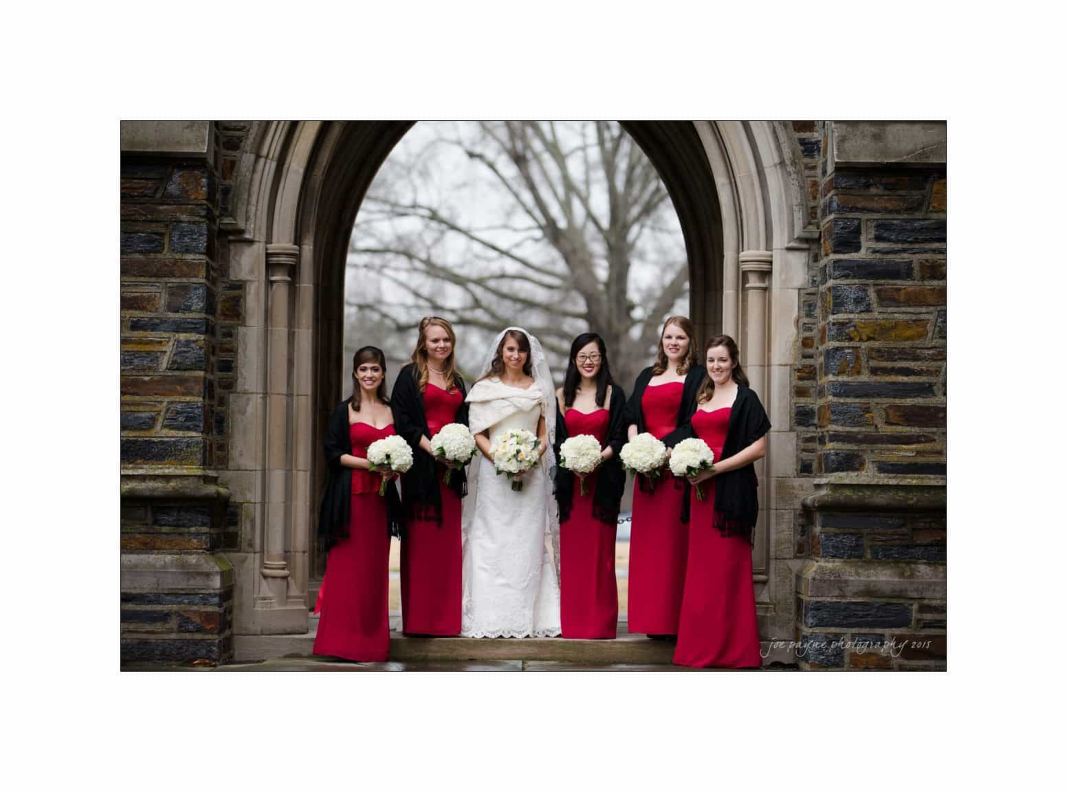 A bride in a white dress stands under a stone archway, surrounded by five bridesmaids wearing matching red dresses and black shawls. Each holds a bouquet of white flowers. Bare trees are visible in the background.