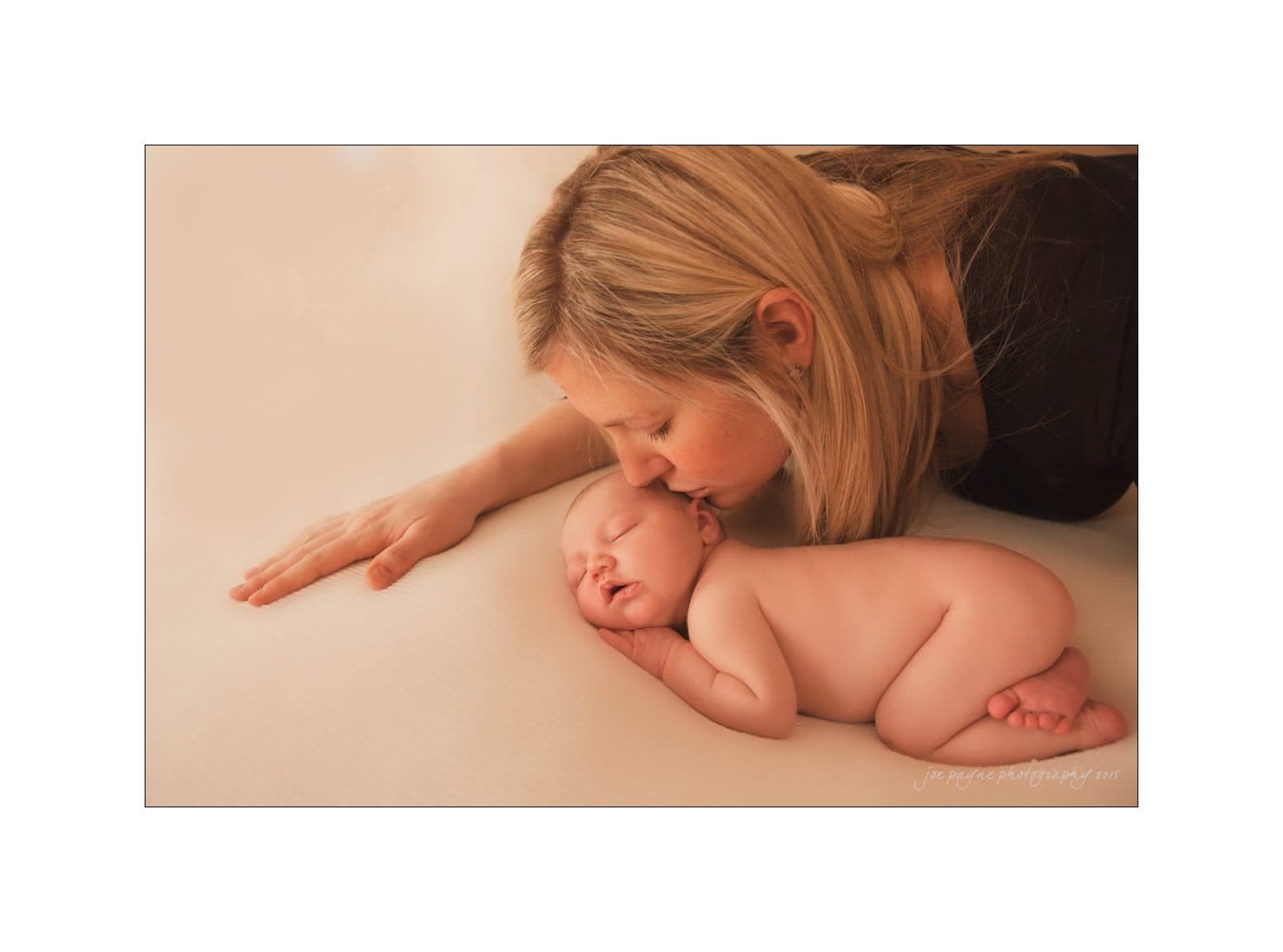 raleigh newborn photographer - baby patton A woman gently kisses the forehead of a sleeping baby lying on a soft surface. Both are positioned closely, conveying tenderness and warmth. The background is a soft, neutral tone.