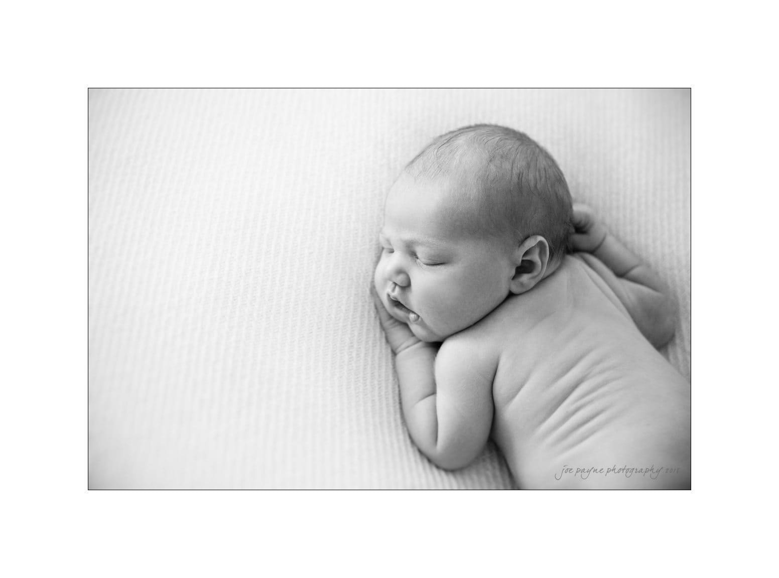 raleigh newborn photographer - baby patton Black and white photo of a newborn baby lying on its stomach on a soft, textured surface. The baby is peacefully sleeping with its head turned to the side, showcasing a serene and gentle expression.