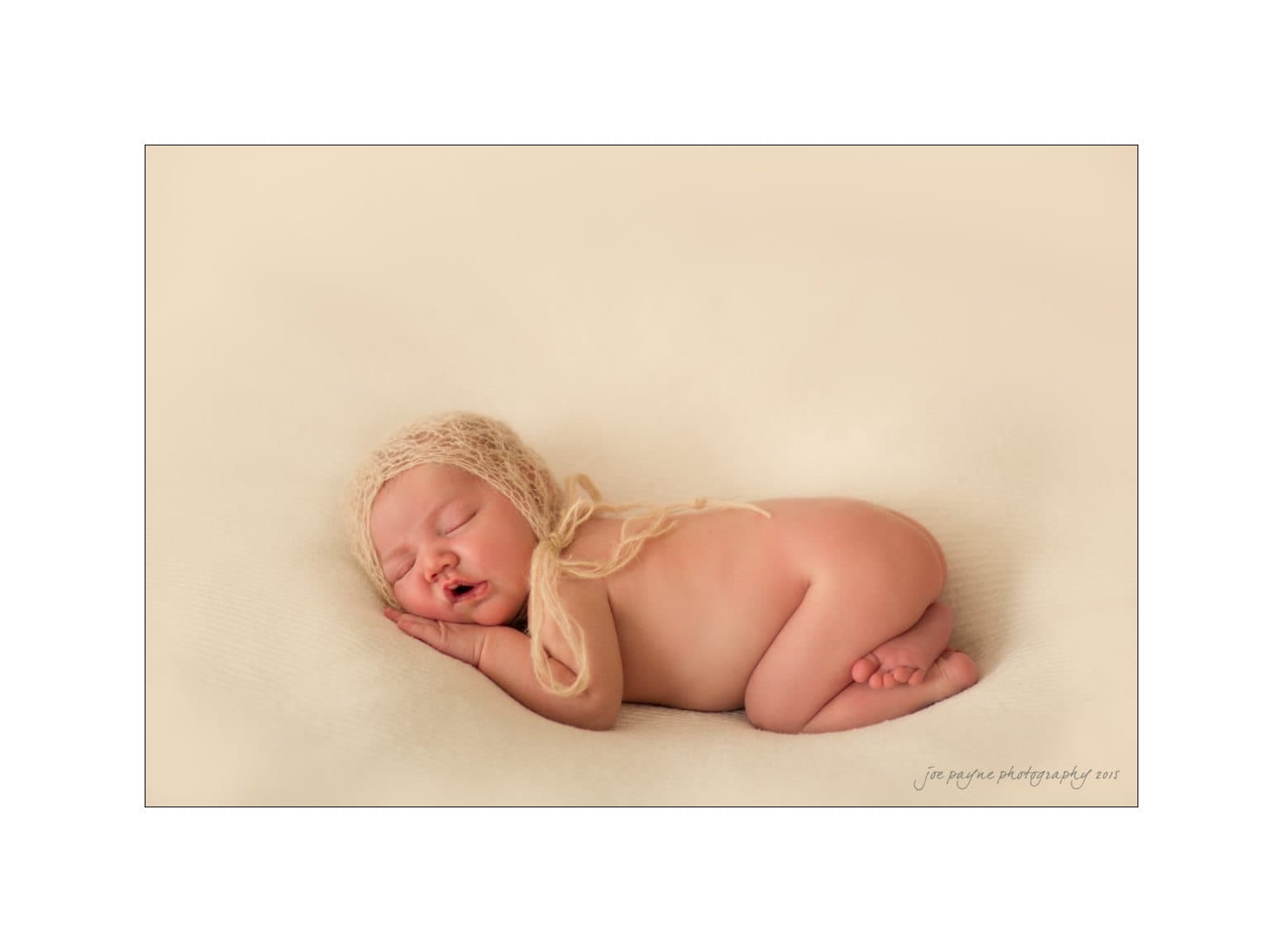 A newborn baby peacefully sleeps on a soft, cream-colored blanket. The baby is wearing a knitted bonnet and is curled up on their side, creating a serene and cozy atmosphere.