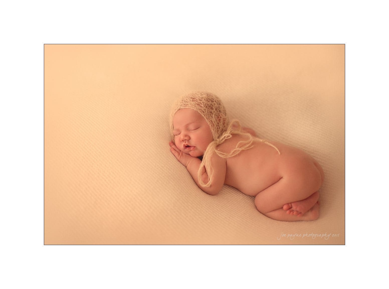 raleigh newborn photographer - baby patton A newborn baby sleeps peacefully on a soft, cream-colored surface. The baby is wearing a delicate, knitted bonnet and is curled up gently, highlighted against the warm-toned background.