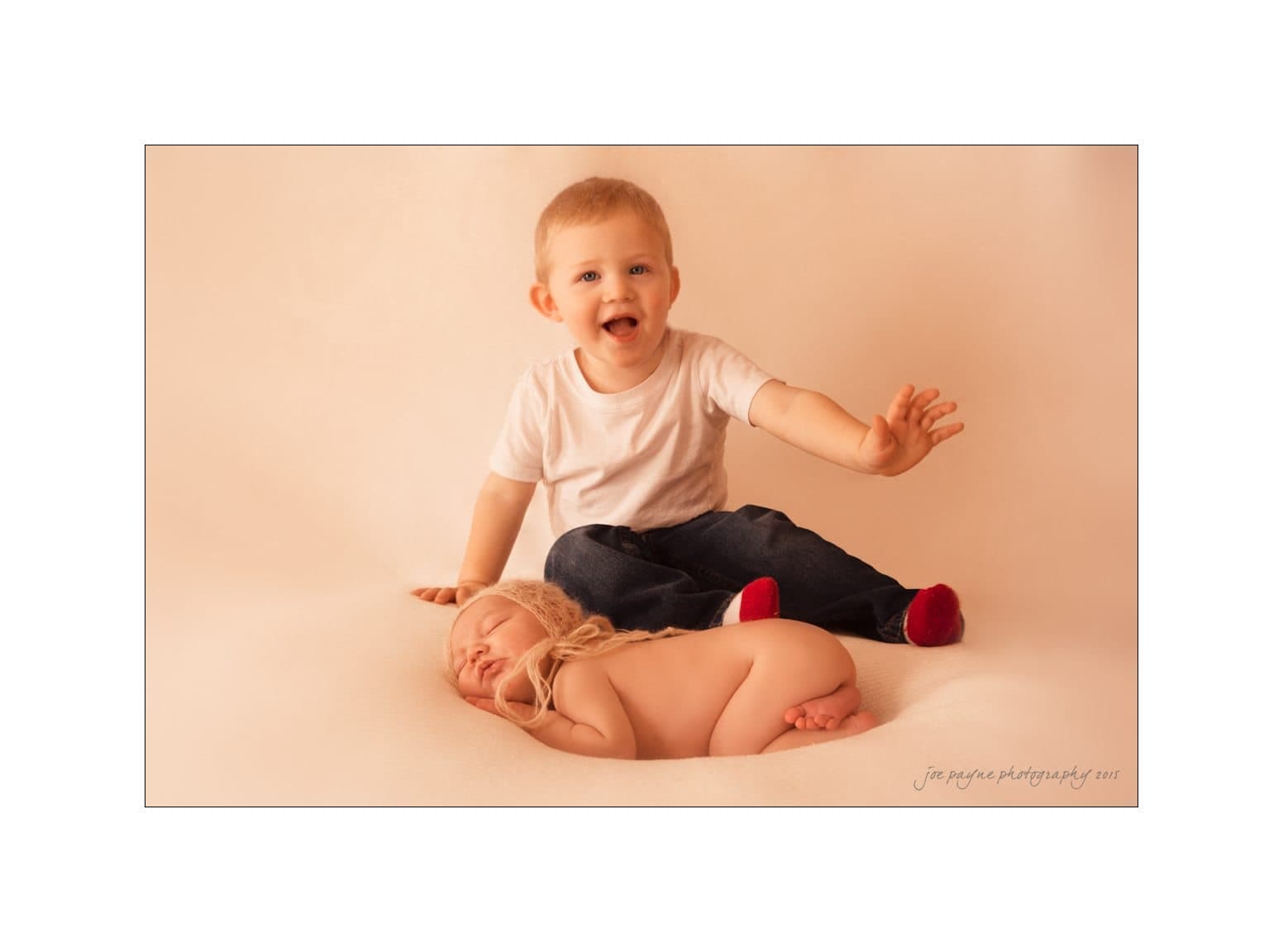 raleigh newborn photographer - baby patton A smiling young boy wearing a white shirt and jeans sits on a soft surface, leaning gently on a sleeping baby nestled in a cozy blanket. The background is a warm beige.