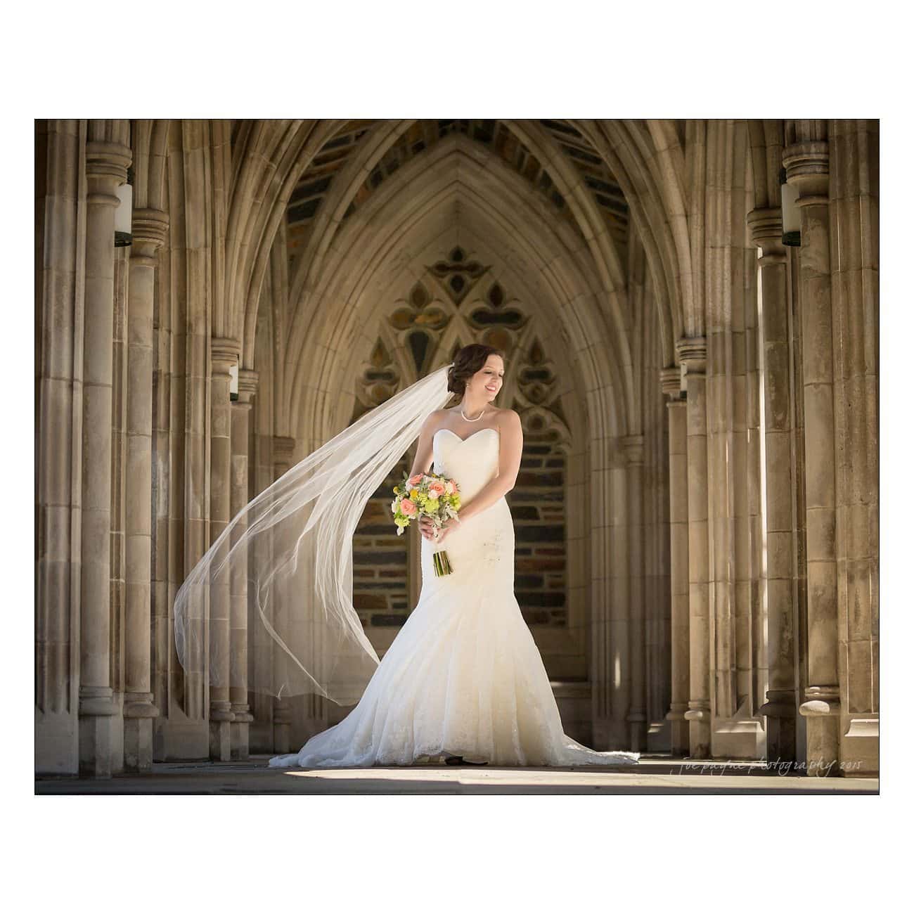 Bride in a strapless gown holds a bouquet, standing in front of a grand, arched stone structure. Her long veil flows elegantly behind her, capturing the light in a serene, picturesque setting.