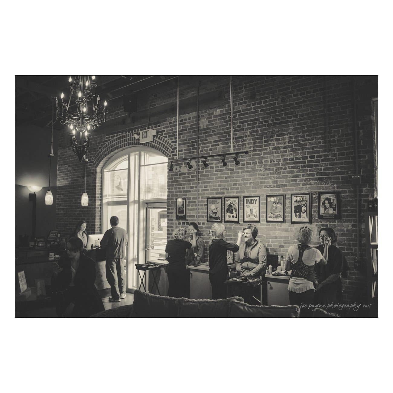A black and white photo of people socializing in a loft with brick walls and large windows. Framed posters line the wall, and a chandelier hangs from the ceiling. Some individuals sit, while others stand and converse.