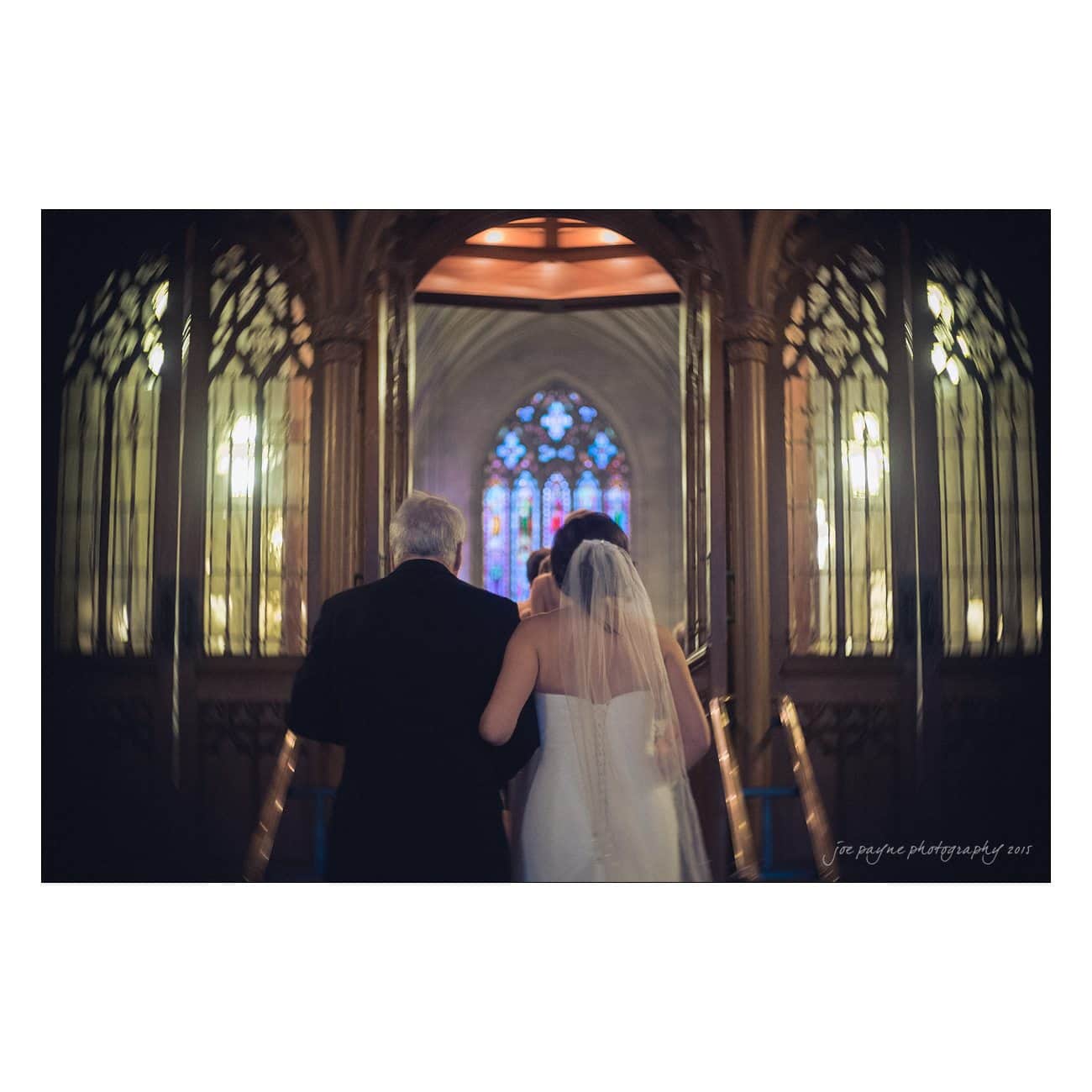 A bride in a white wedding dress and veil walks down the aisle with an older man in a suit. Theyre in a church with stained glass windows and soft lighting.