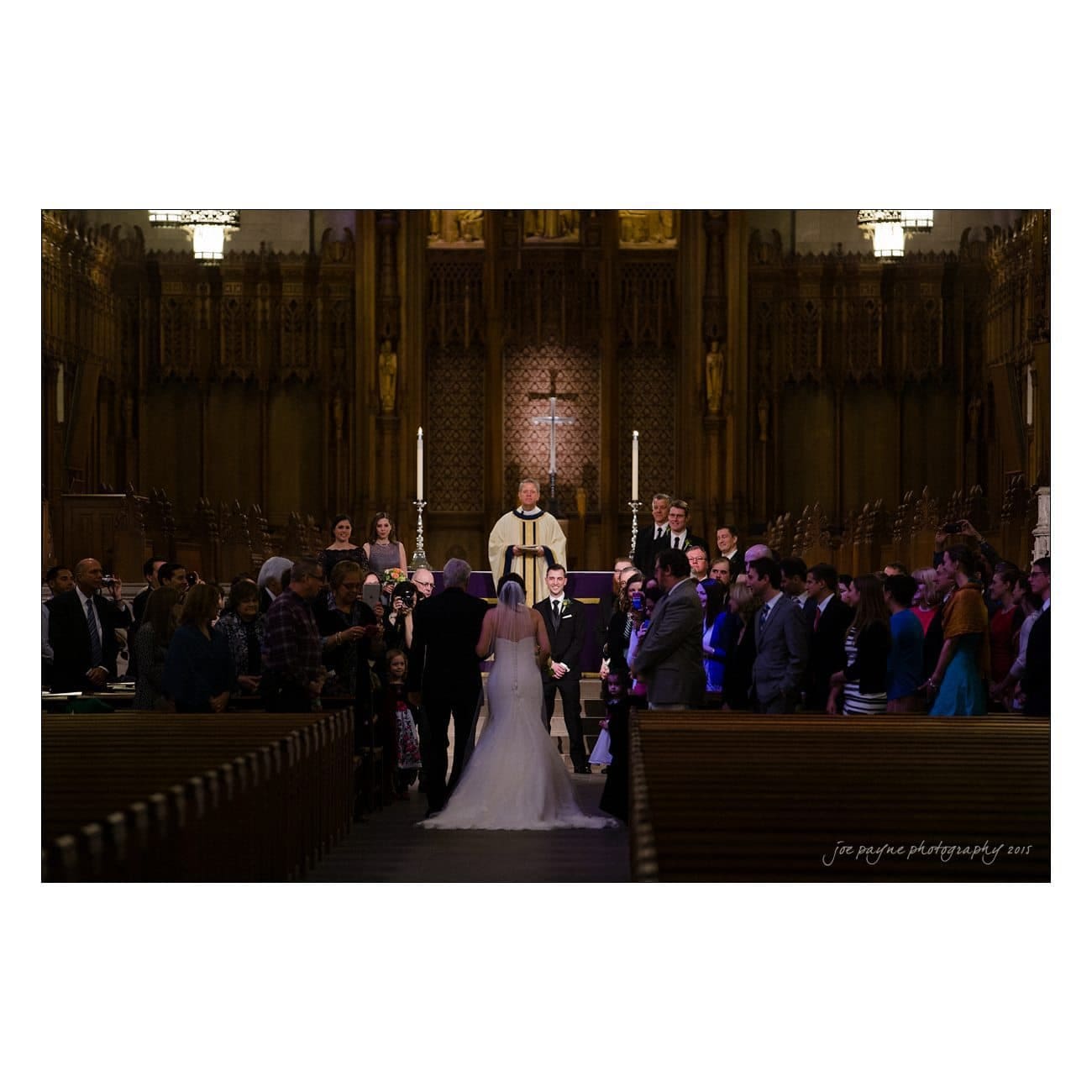 A bride and groom stand at the altar in a dimly lit church, surrounded by friends and family. The officiant is in the center, and the guests are seated in pews on both sides, facing the couple. A large cross is visible in the background.