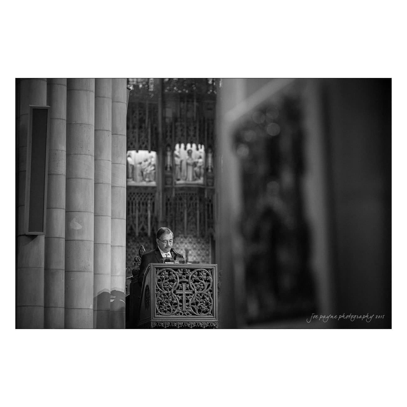 A man stands at an ornate pulpit in a cathedral, reading from a document. The background features intricate stone carvings and columns. The scene is in black and white, highlighting architectural details.