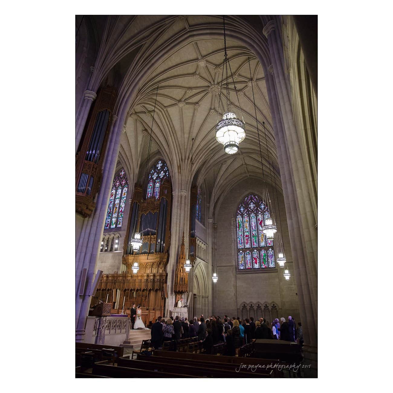 A grand cathedral interior with high vaulted ceilings, stained glass windows, and a large chandelier. A wedding scene is unfolding with guests seated, and a couple standing at the altar near a wooden organ.