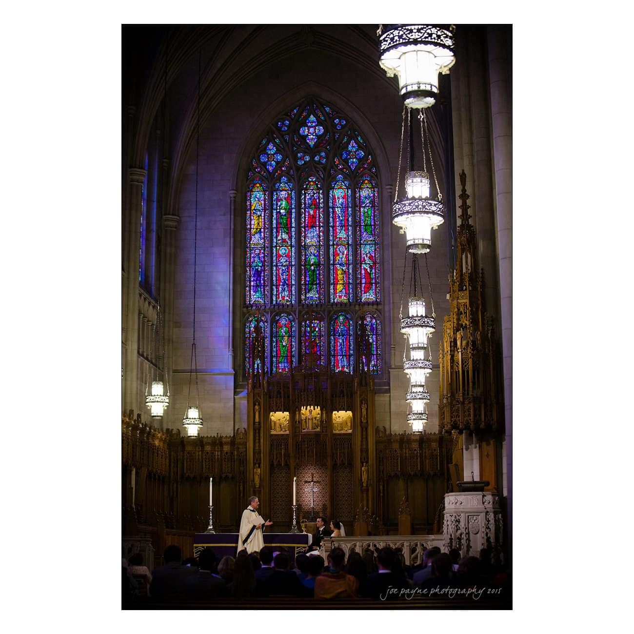 A church interior with arched stained glass windows, ornate wooden carvings, and hanging chandeliers. A person in white robes stands near the altar, addressing attendees seated in pews.