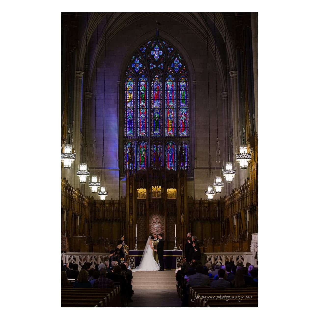A couple stands at the altar during a wedding ceremony in a grand church with stained glass windows and high vaulted ceilings. They are surrounded by the wedding party, guests seated in pews, and lit by ornate chandeliers.