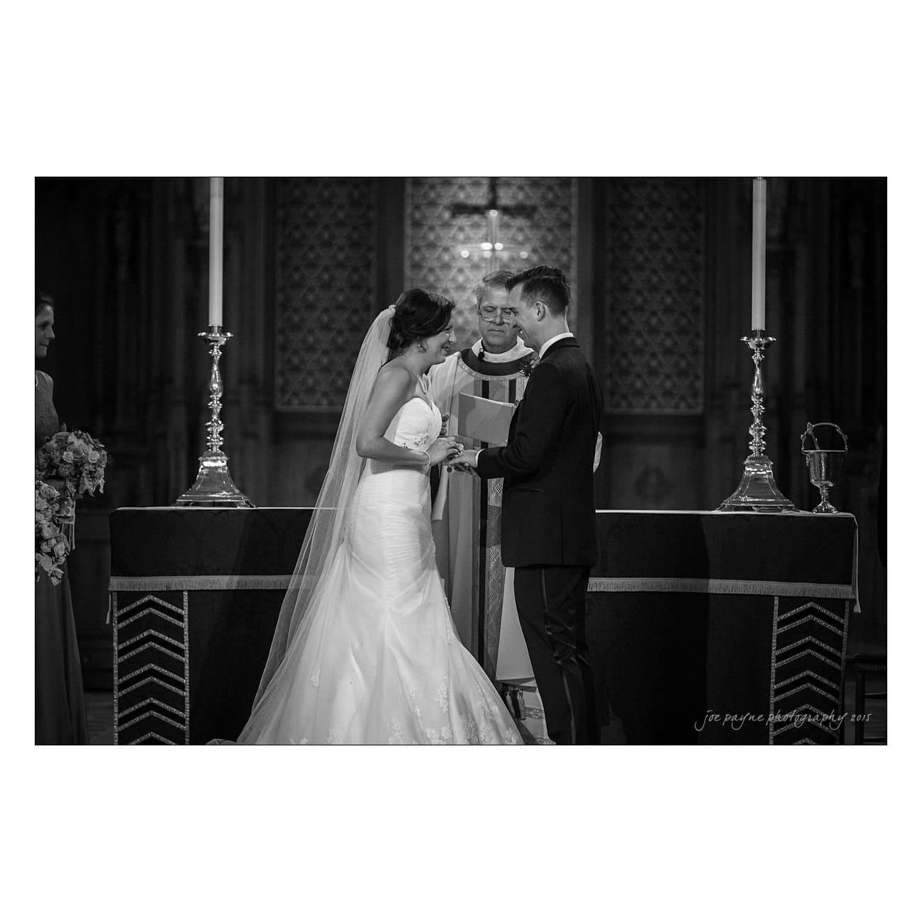 A bride and groom stand at the altar during a wedding ceremony, holding hands. A priest stands between them, officiating. The setting is a church with decorative candles and religious symbols in the background.