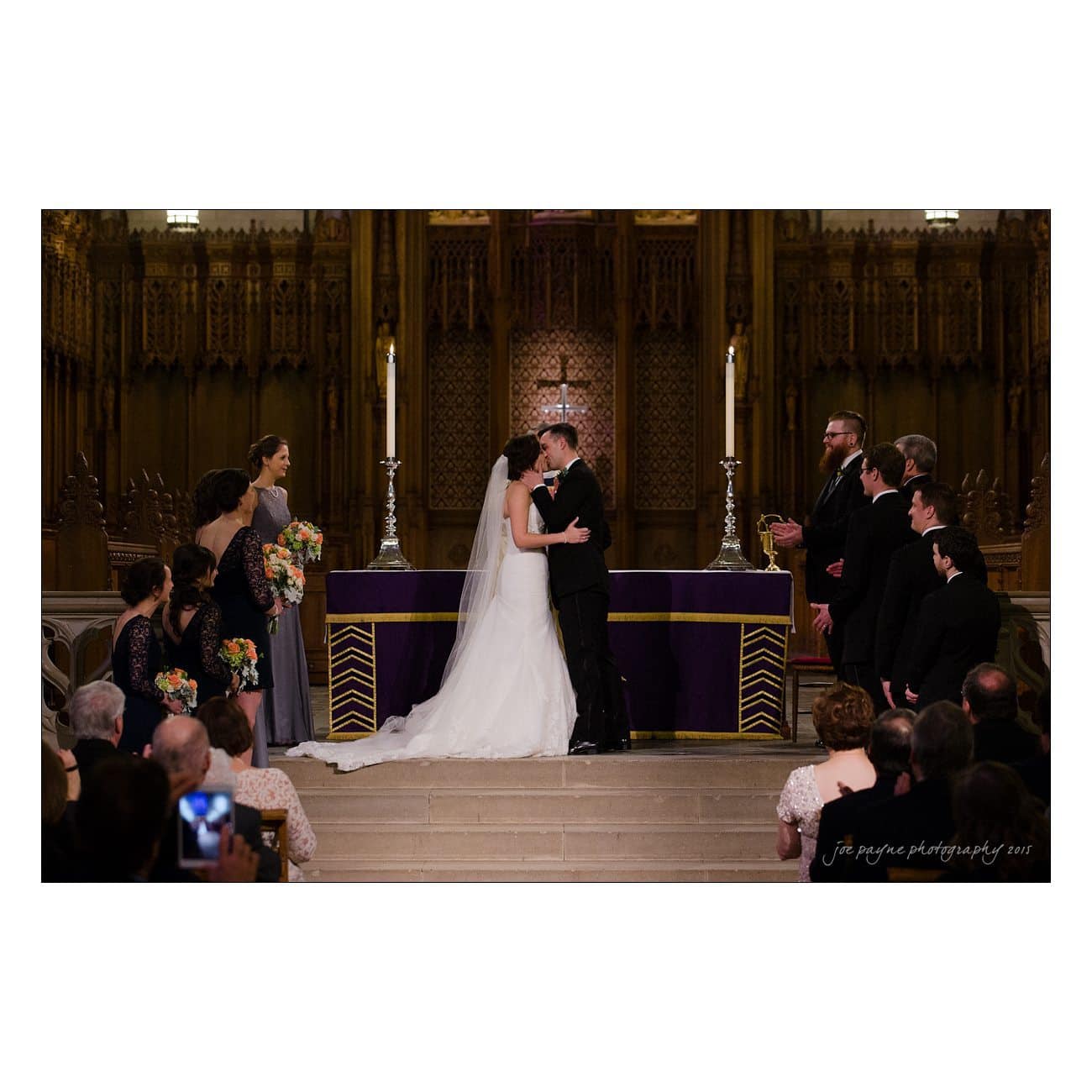A bride and groom share a kiss at the altar during a wedding ceremony inside a church. Bridesmaids and groomsmen stand on either side, and guests are seated in the foreground. Lit candles and ornate decorations are visible in the background.