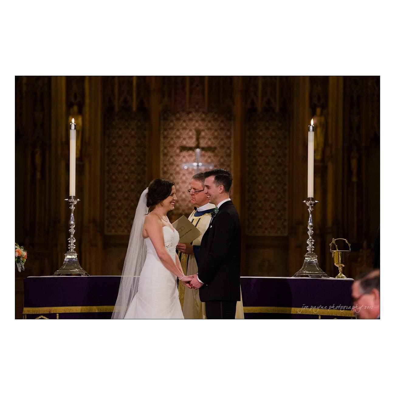 A bride and groom stand holding hands during their wedding ceremony in a church. The officiant, dressed in religious attire, stands between them. Candles and a cross are visible in the background.